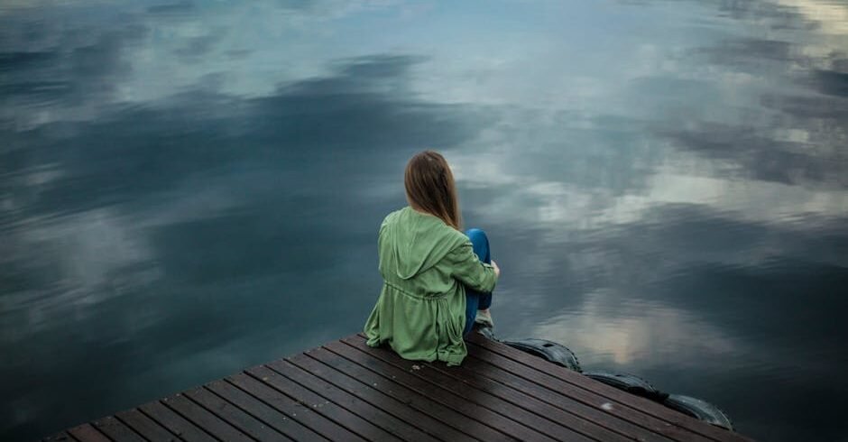 A woman sits on a wooden dock, reflecting by a calm lake under a cloudy sky.