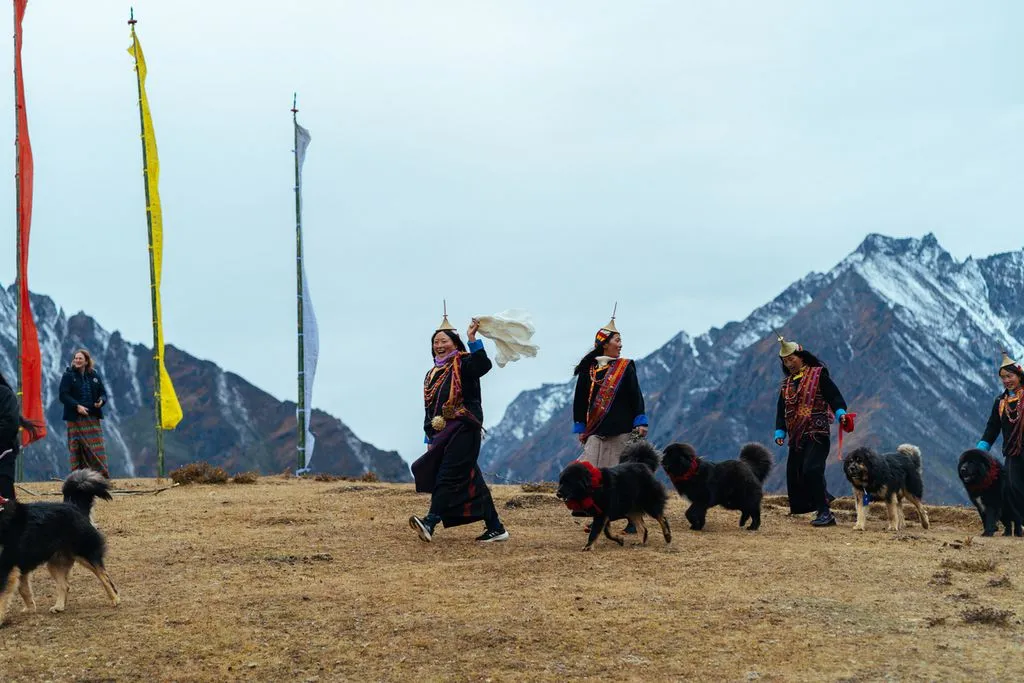 Layap women in traditional dress and conical bamboo hats walking with their dogs below prayer flags and snow-capped peaks in Laya