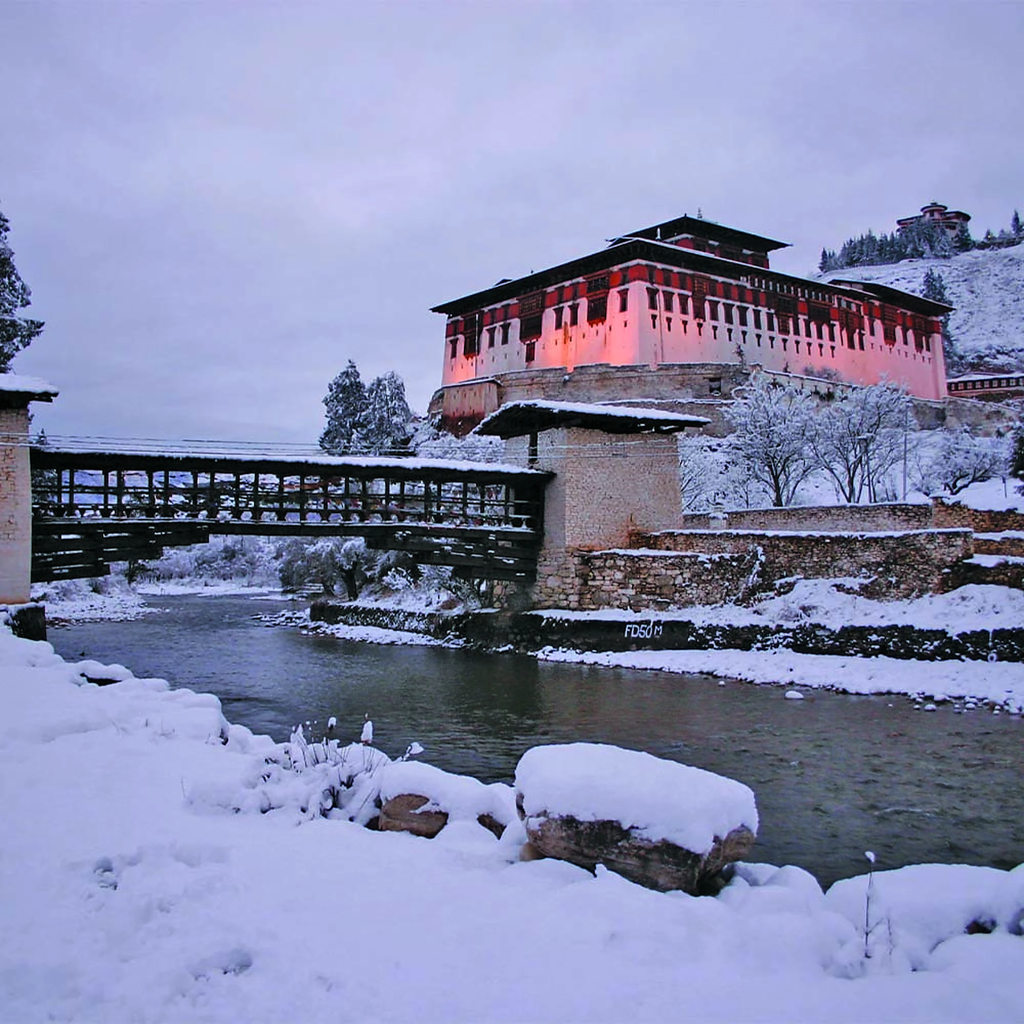 Rinpung Dzong and its covered wooden bridge over the Paro Chhu after snowfall, with the Ta Dzong on the hill above